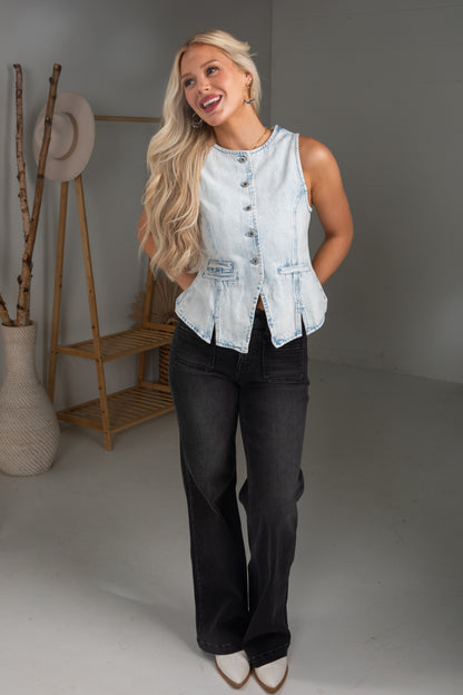 Woman wearing a light blue denim vest over a black top and jeans, standing in a minimalistic room.