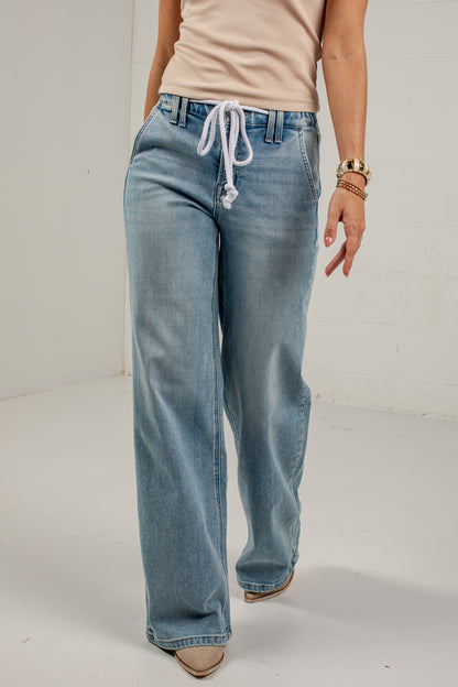 A person wearing light-wash blue wide-leg jeans with a white drawstring waist, paired with a beige top and beige shoes, standing against a plain white studio background.