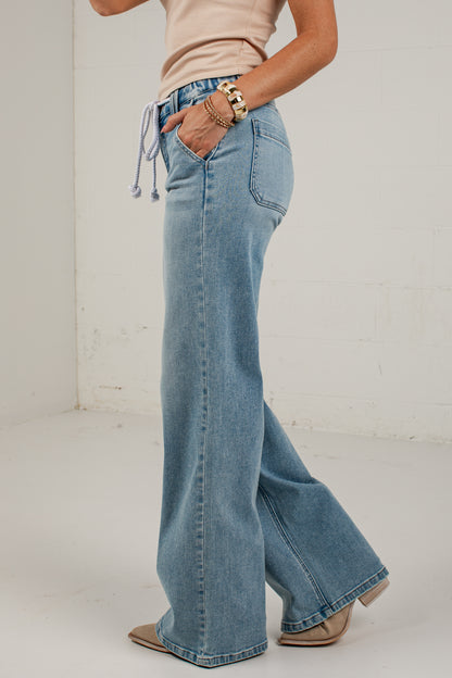 A person wearing light blue wide-leg denim jeans with a drawstring waist, paired with a beige top and beige shoes, standing in a white studio corner.