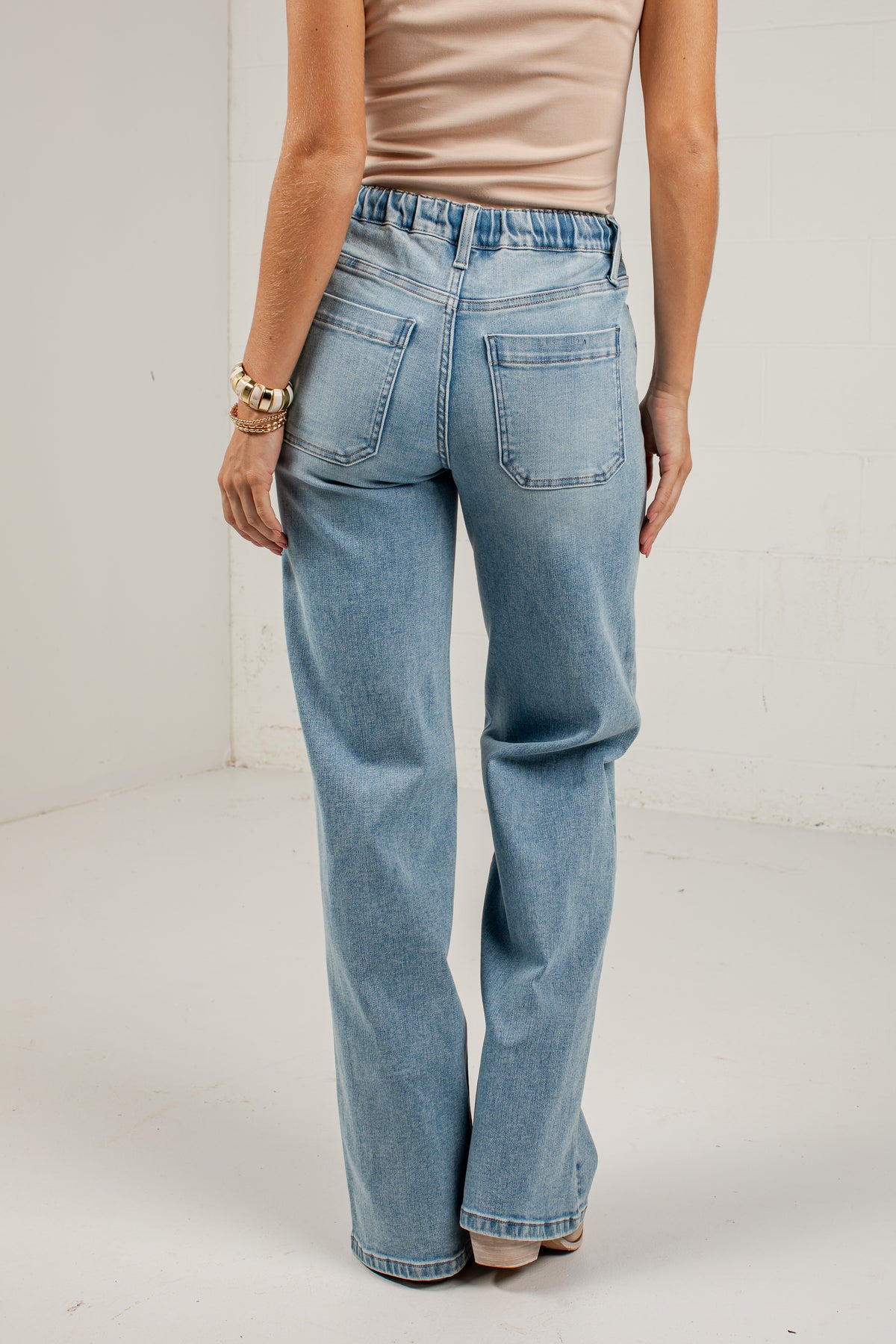 A model shown from the back wearing light-wash blue wide-leg jeans, a beige ribbed tank top, and beige heels, standing in a white studio corner.