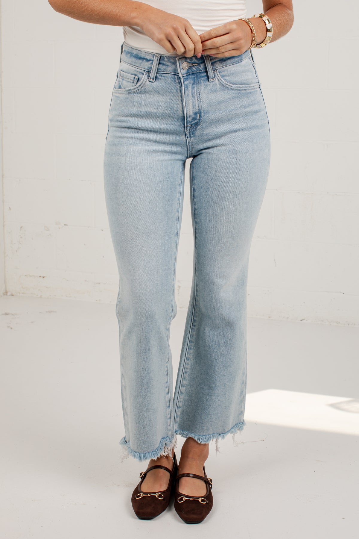 A person wearing light-wash blue cropped flare jeans with a raw frayed hem, paired with a white top and dark brown flat shoes, standing against a white wall.