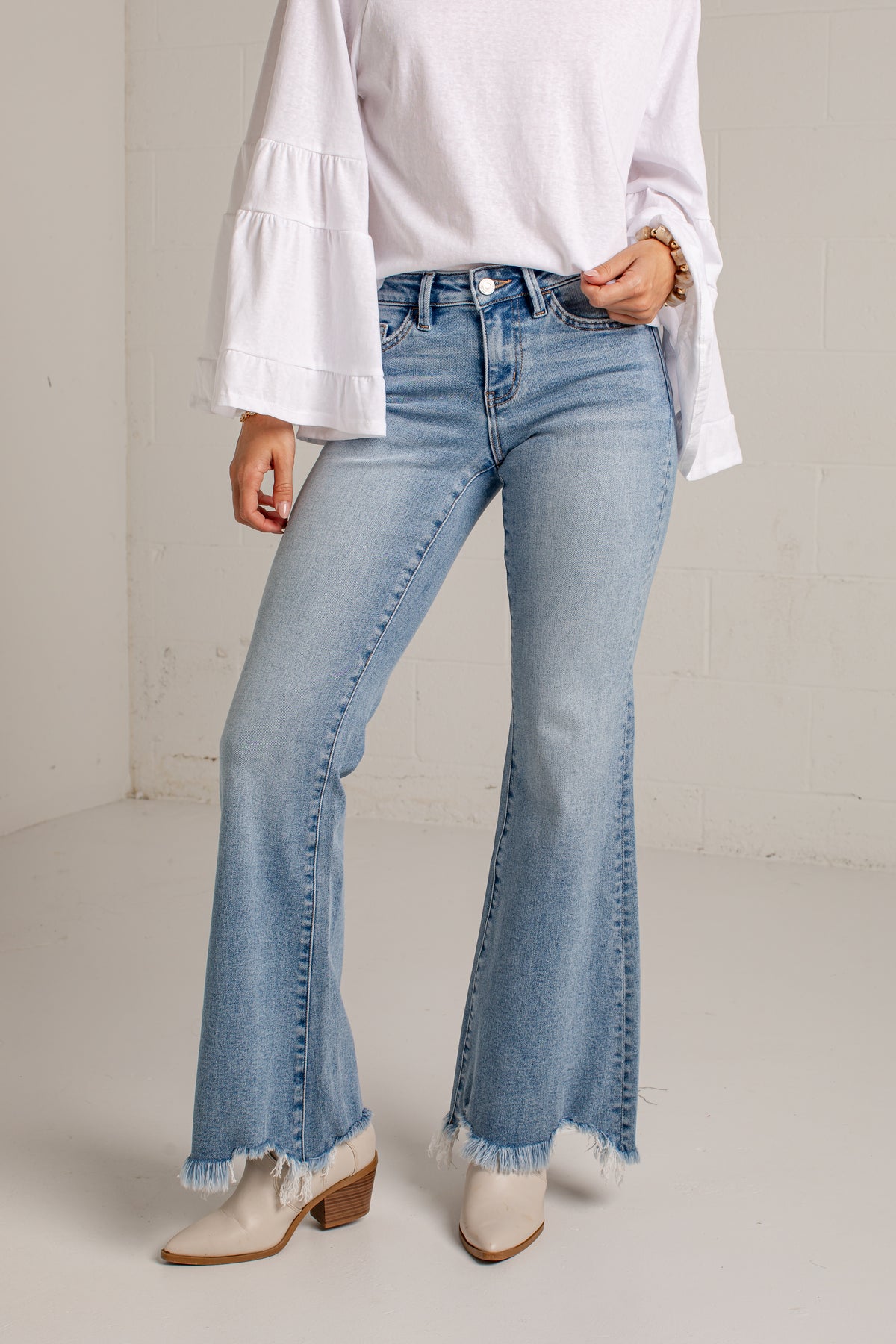 A model wearing light-wash blue flare jeans with frayed hems, paired with a white long-sleeve blouse and white heeled boots, standing against a light gray brick wall.