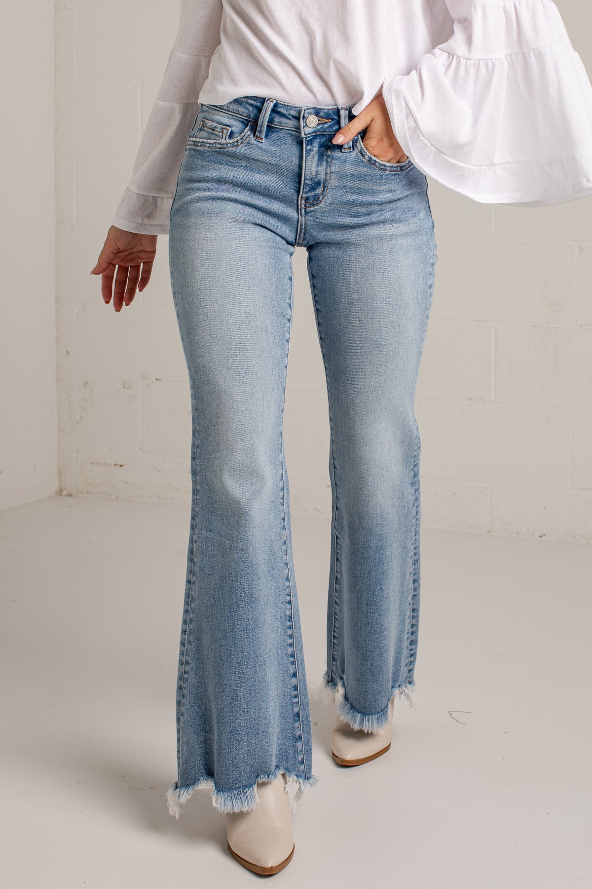 A person wearing light-wash blue flare jeans with frayed hems, paired with a white blouse and white pointed-toe heels, standing against a light neutral wall.