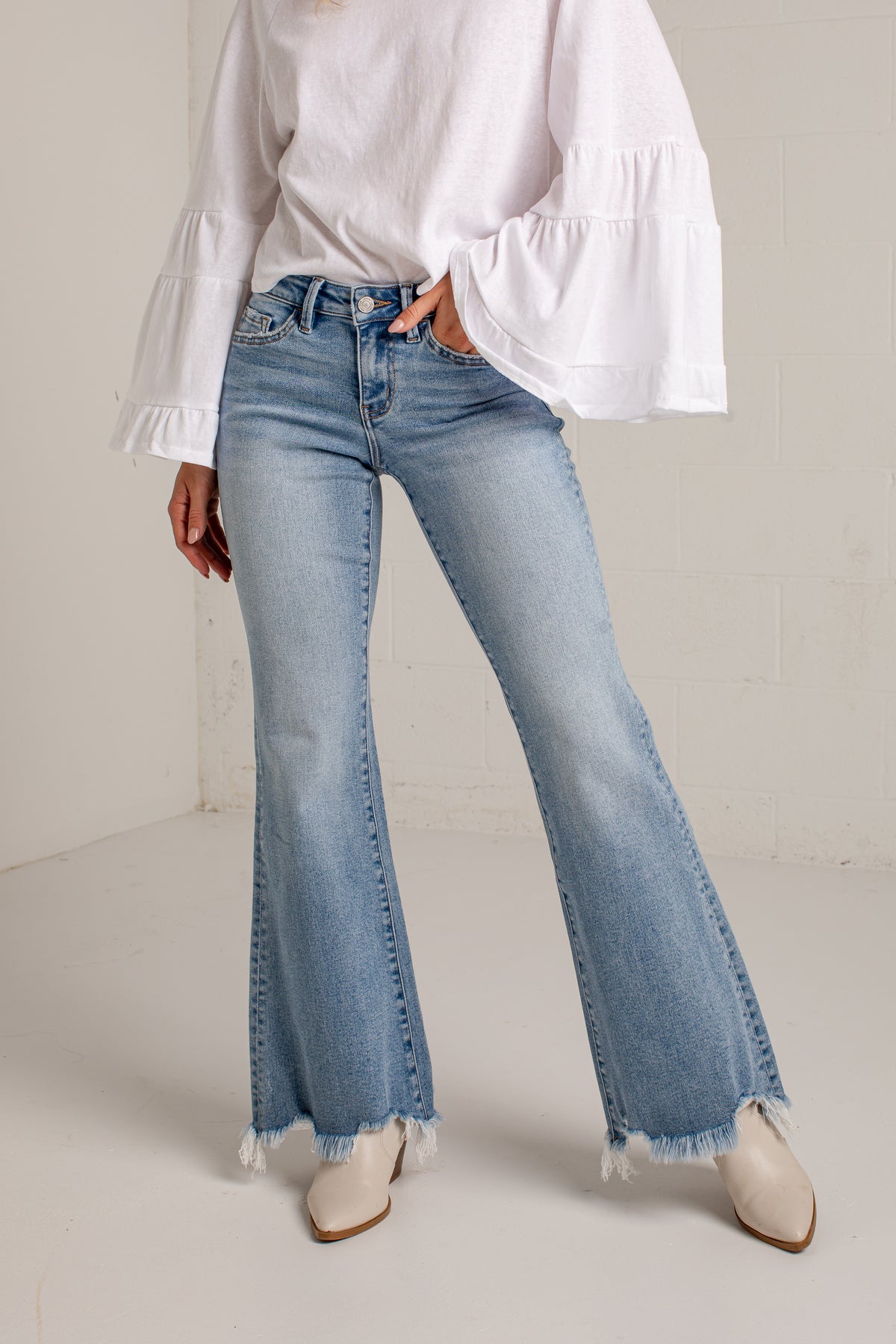 A person wearing light-wash blue flare jeans with frayed hems, paired with a white tiered blouse and beige pointed-toe boots, standing against a light neutral wall.