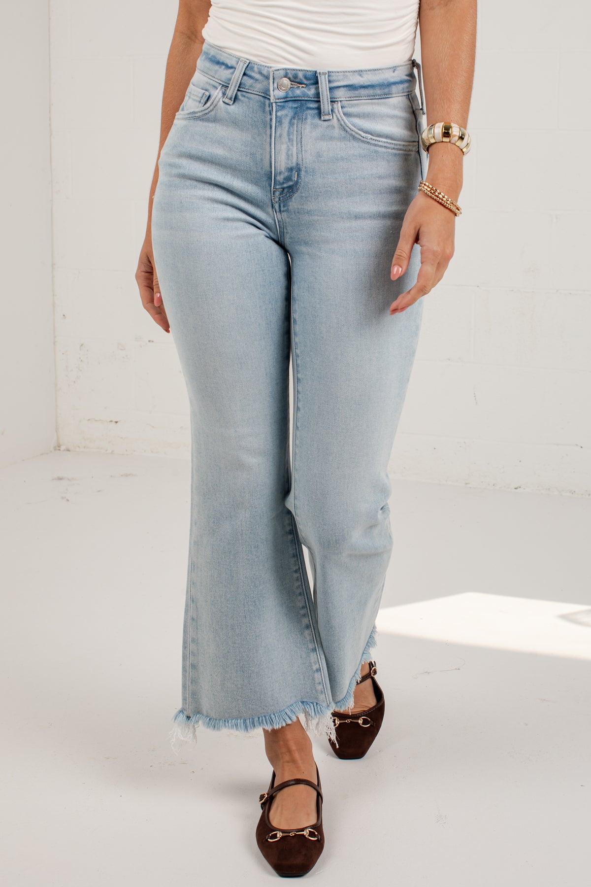 A person wearing light-wash blue cropped flare jeans with a raw frayed hem, paired with a white top and dark brown flat shoes, standing against a white studio wall.
