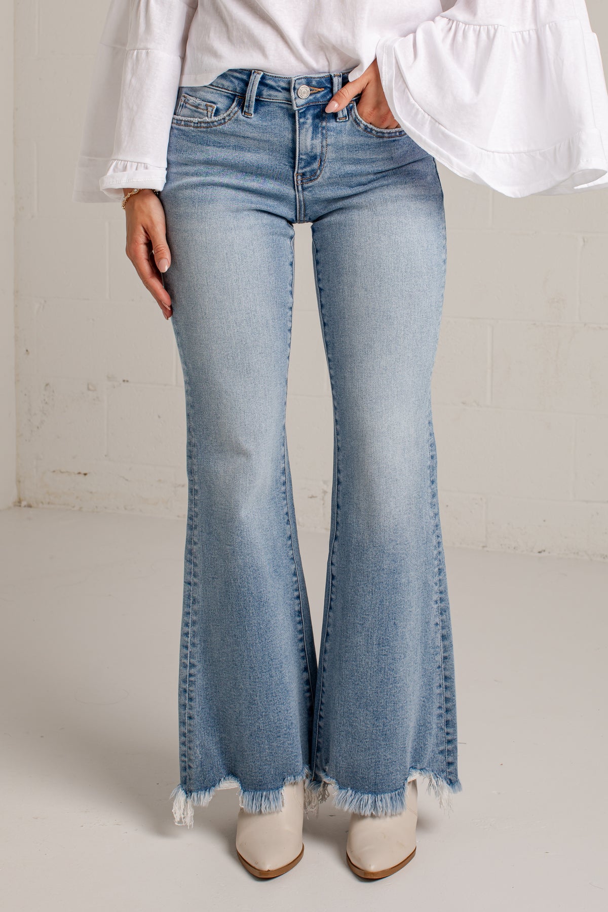 A person wearing light-wash blue flare jeans with a frayed hem, paired with a white blouse and white pointed-toe heels, standing against a light neutral wall.