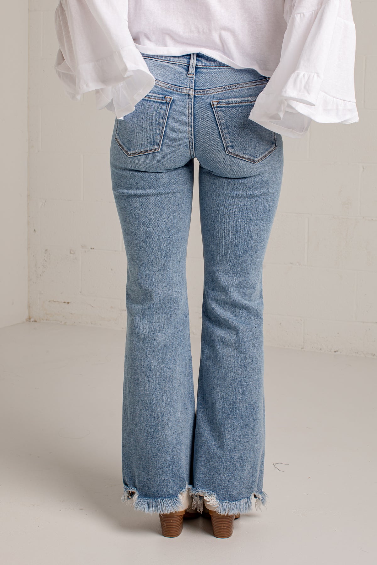 Rear view of a person wearing light-wash blue flare jeans with a frayed hem, paired with a white blouse and brown heels, standing against a plain off-white wall.