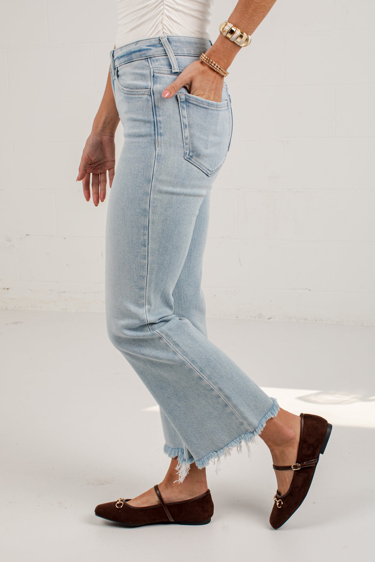 A person wearing light-wash cropped denim jeans with a raw frayed hem, paired with dark brown flat shoes and gold jewelry, standing against a white studio background.