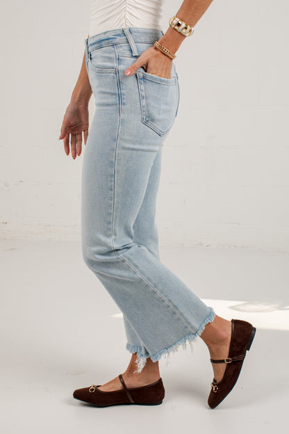 A person wearing light-wash cropped denim jeans with a raw frayed hem, paired with dark brown flat shoes and gold jewelry, standing against a white studio background.