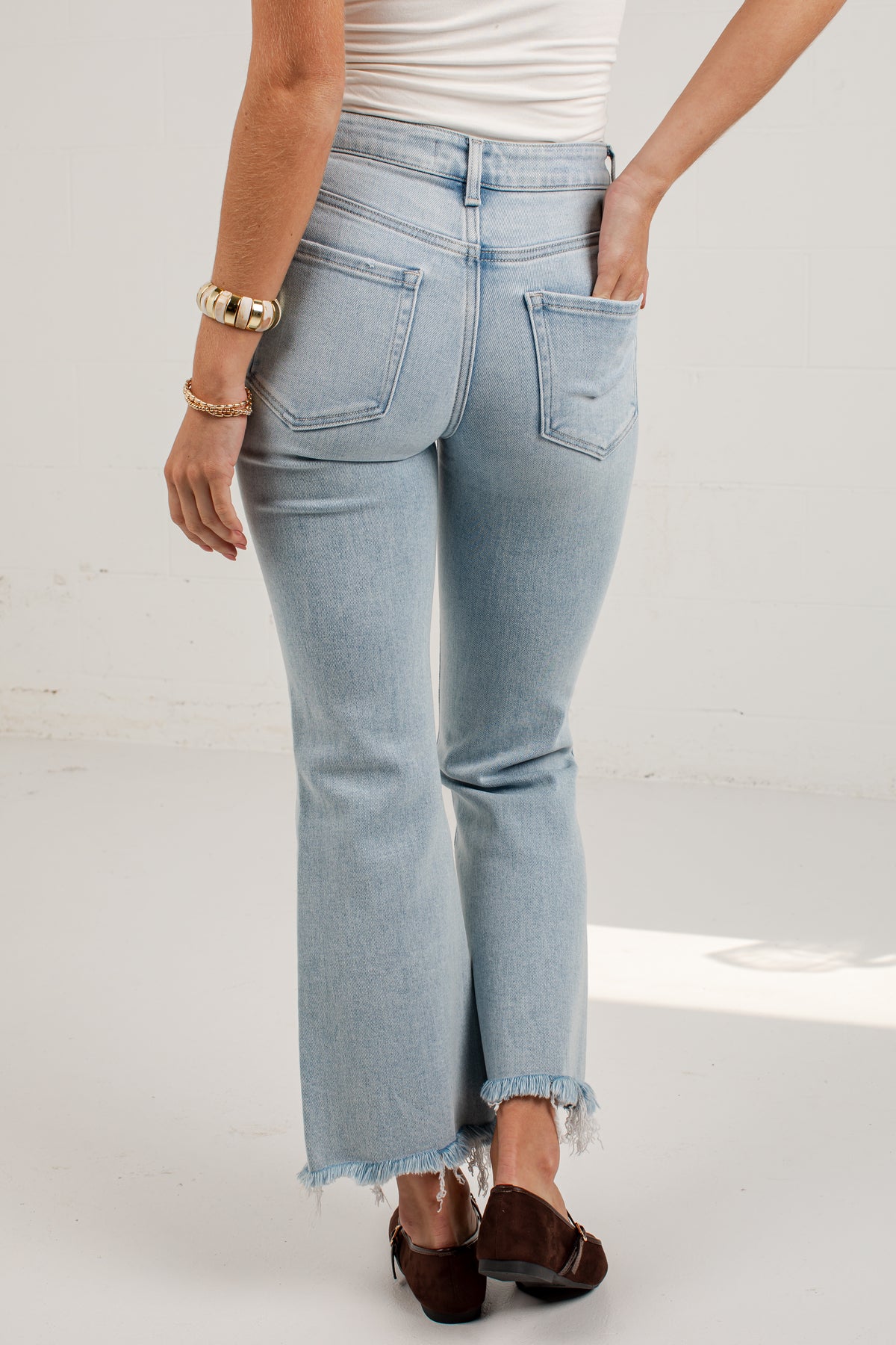 A person shown from the back wearing light-wash blue cropped jeans with a raw frayed hem, paired with a white top and dark brown flats, against a plain light studio background.