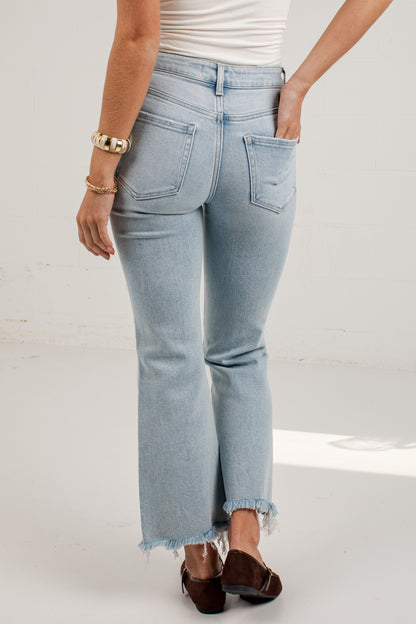 A person shown from the back wearing light-wash blue cropped jeans with a raw frayed hem, paired with a white top and dark brown flats, against a plain light studio background.
