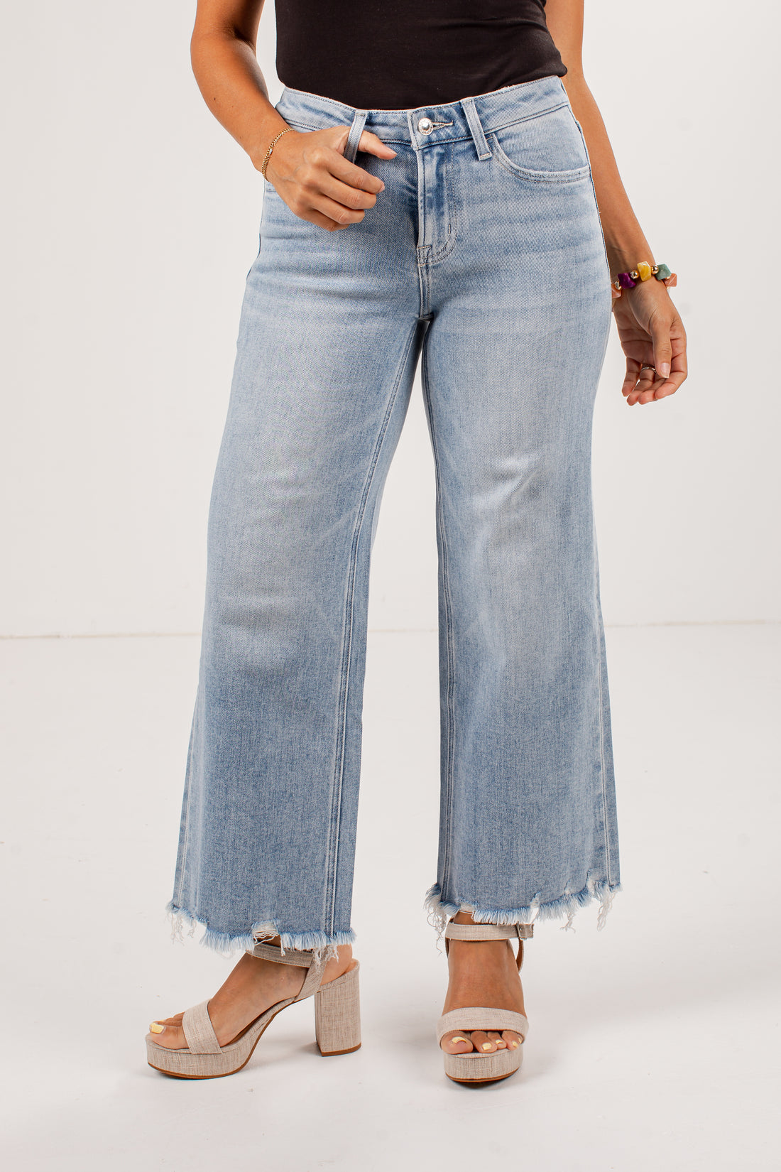 A model wearing light-wash blue wide-leg cropped jeans with a raw frayed hem, paired with beige platform sandals and a black top, shown against a plain white studio background.
