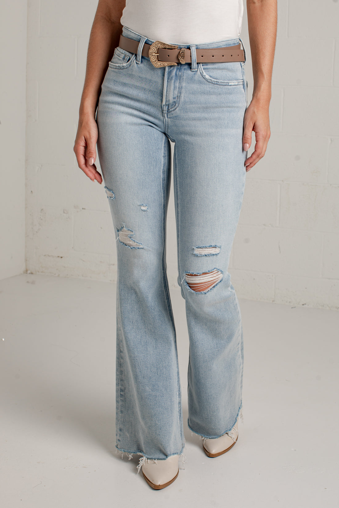A person wearing light-wash blue distressed flare jeans with a brown belt, standing against a white studio wall.