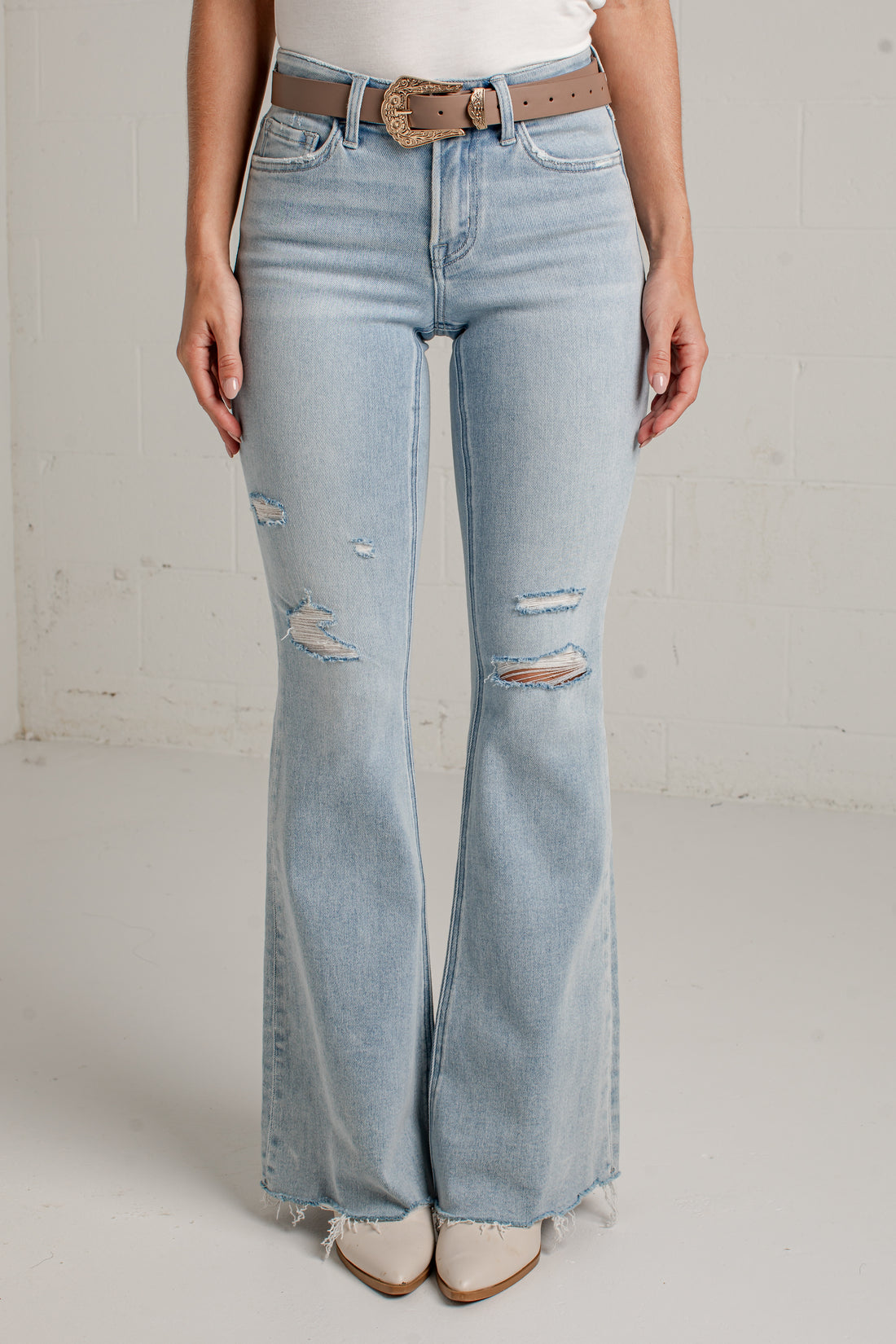 A person wearing light-wash blue distressed flare jeans with frayed hems, paired with a tan belt and white pointed-toe shoes, standing against a white brick wall.