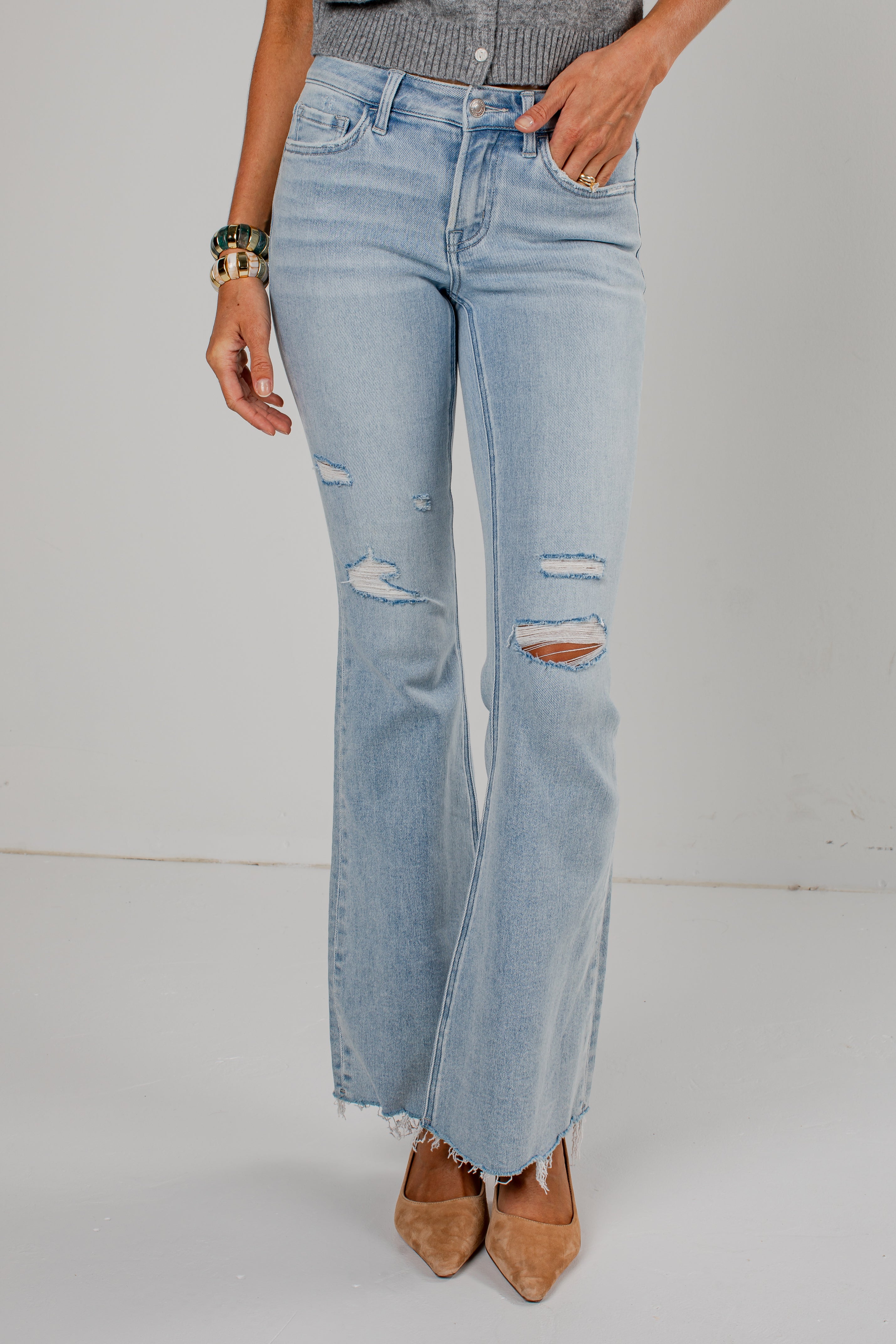 A person wearing light-wash blue distressed flare jeans with frayed hems, paired with tan pointed-toe heels and a gray top, standing against a plain light gray studio background.