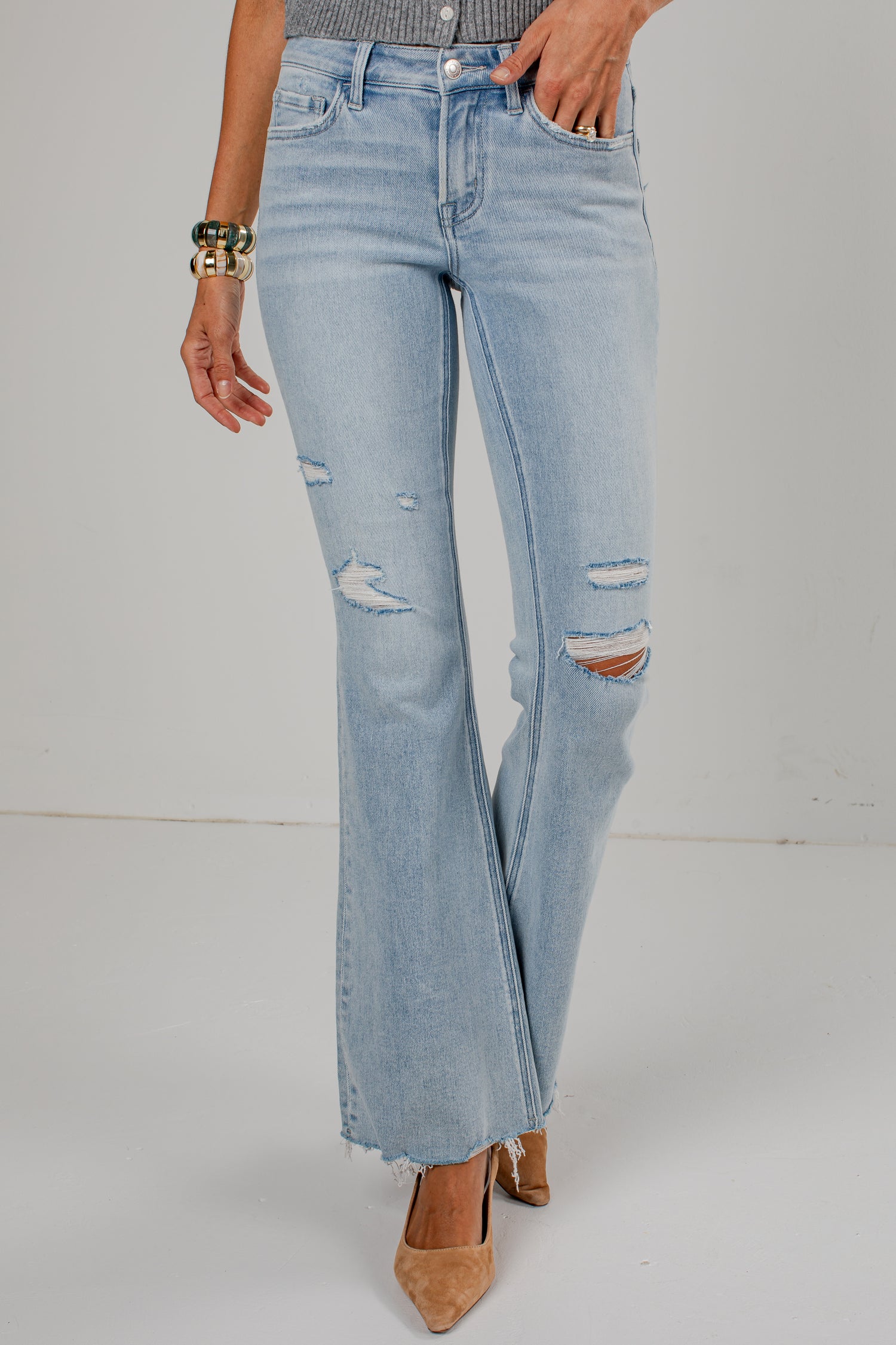 Light-wash blue distressed flare jeans with ripped details on the thighs and knees, shown on a model wearing beige pointed-toe heels against a white studio background.