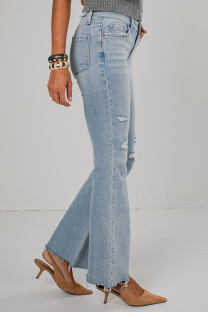 A person wearing light-wash blue distressed flare jeans with frayed hems, paired with tan pointed-toe heeled mules and bracelets, photographed against a plain light gray studio background.
