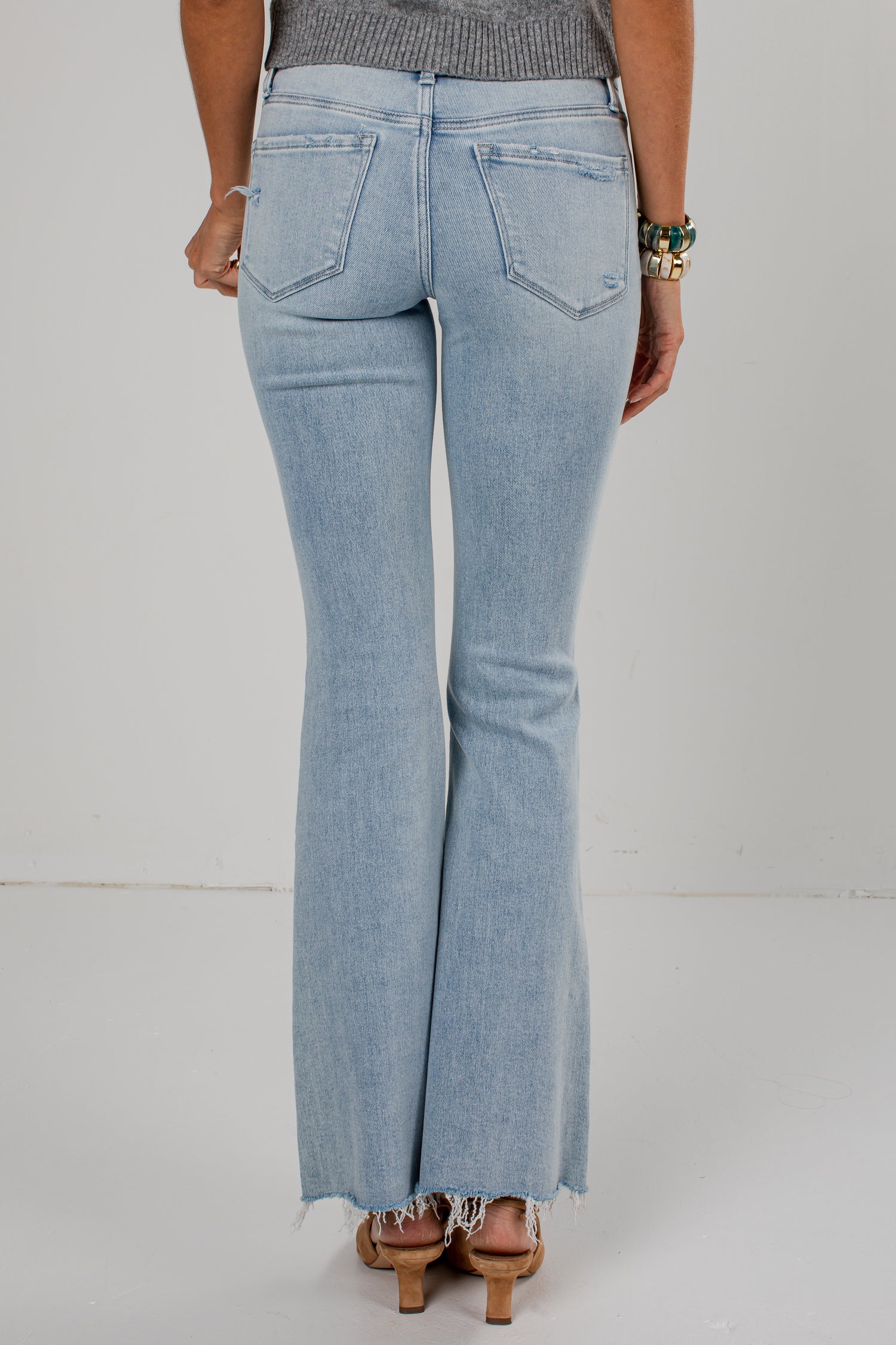 Rear view of a person wearing light-wash blue flare jeans with a frayed hem, paired with a gray top and heeled sandals, against a plain light background.