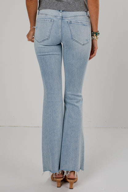 Rear view of a person wearing light-wash blue flare jeans with a frayed hem, paired with a gray top and heeled sandals, against a plain light background.