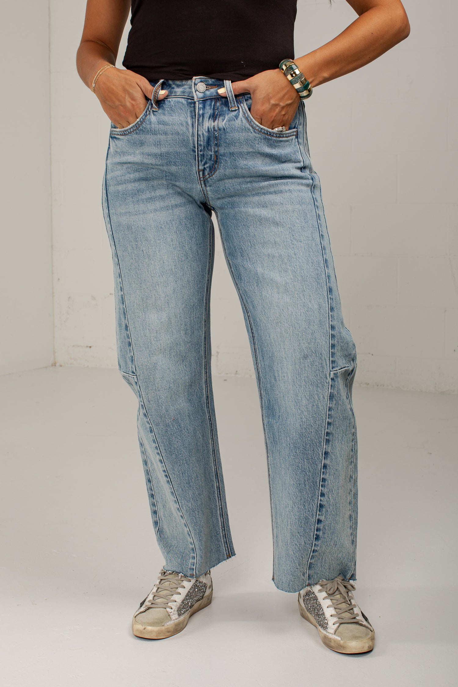 A person wearing light-wash blue straight-leg jeans with side cargo pockets, paired with a black top and distressed sneakers, standing against a plain light gray wall.
