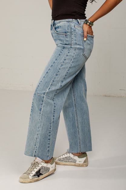 A person wearing light-wash wide-leg denim jeans, a black top, and distressed sneakers, standing against a plain light gray studio wall.