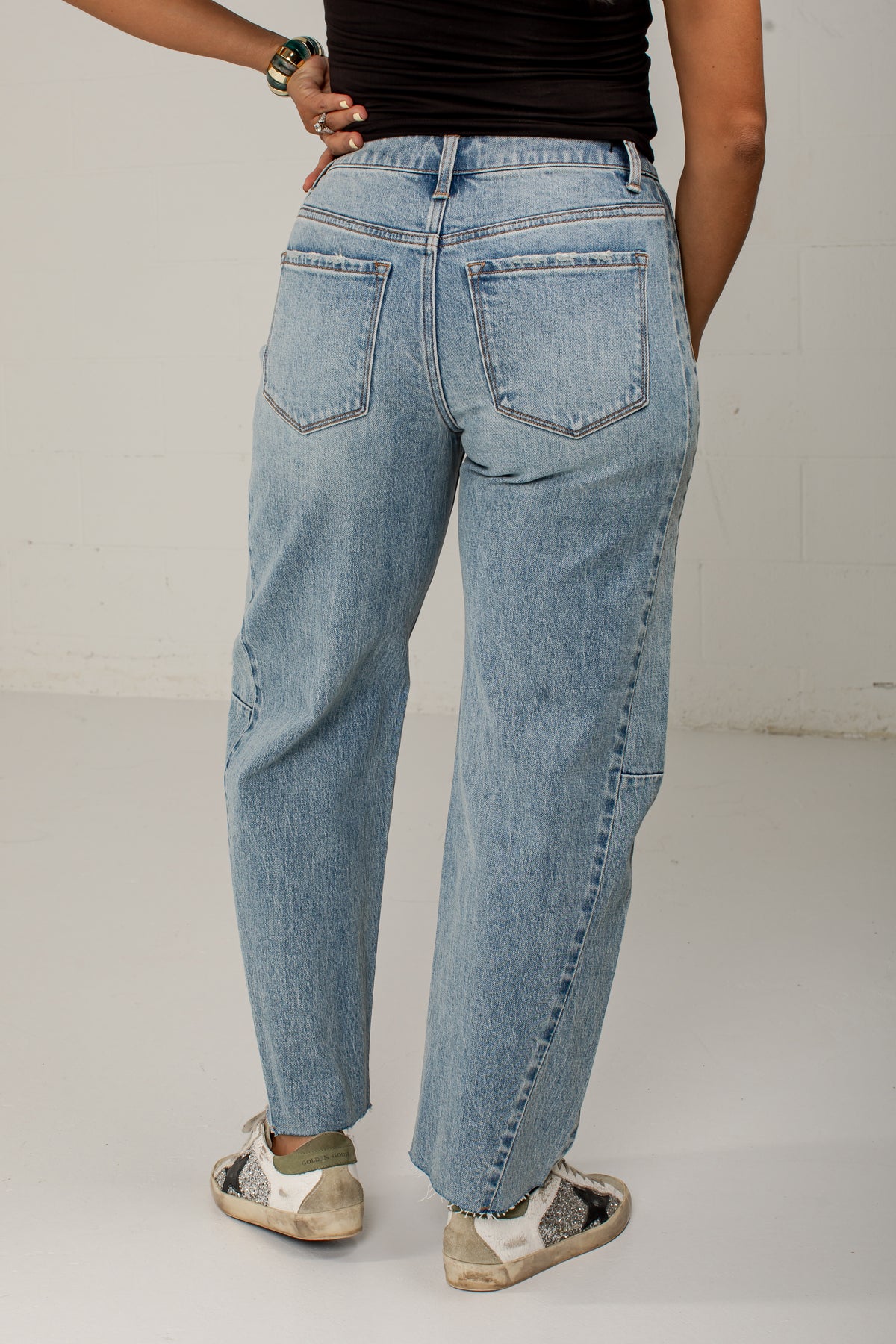 A person shown from the back wearing light-wash blue denim jeans with a straight-leg or relaxed fit, paired with a black top and white sneakers, standing against a plain light wall.