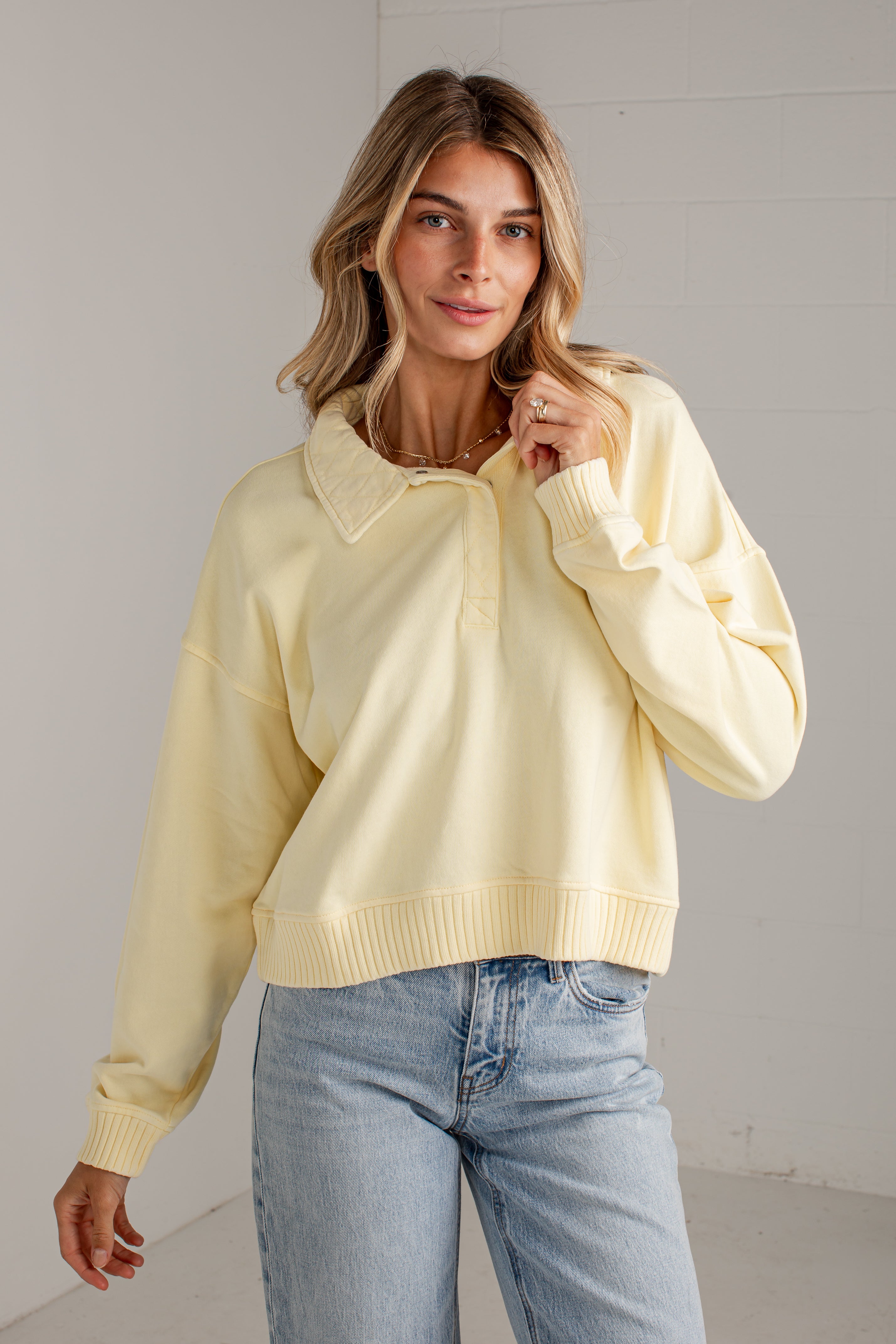 A woman wearing a pale yellow long-sleeve polo-style sweater with a ribbed collar, placket, and hem, paired with light blue jeans, standing against a light gray wall.