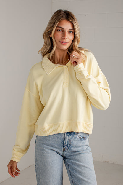 A woman wearing a pale yellow long-sleeve polo-style sweater with a ribbed collar, placket, and hem, paired with light blue jeans, standing against a light gray wall.