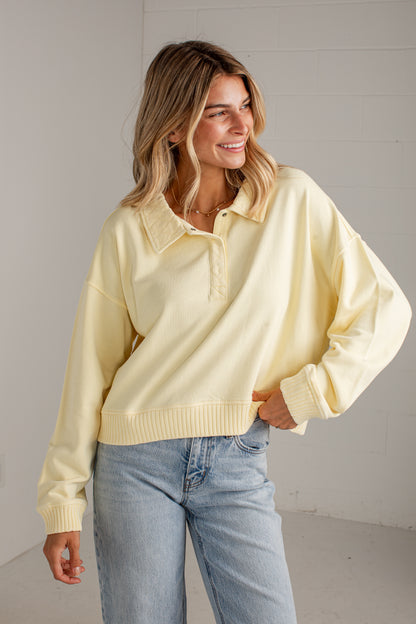 A woman wearing a pale yellow long-sleeve knit polo-style sweater with a ribbed collar, placket, and cuffs, paired with light blue jeans, standing against a light gray wall.