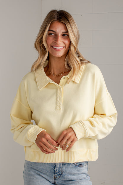 A woman wearing a pale yellow knit polo sweater with a ribbed collar, half-placket, and ribbed cuffs and hem, paired with light blue jeans, standing against a light gray brick wall.
