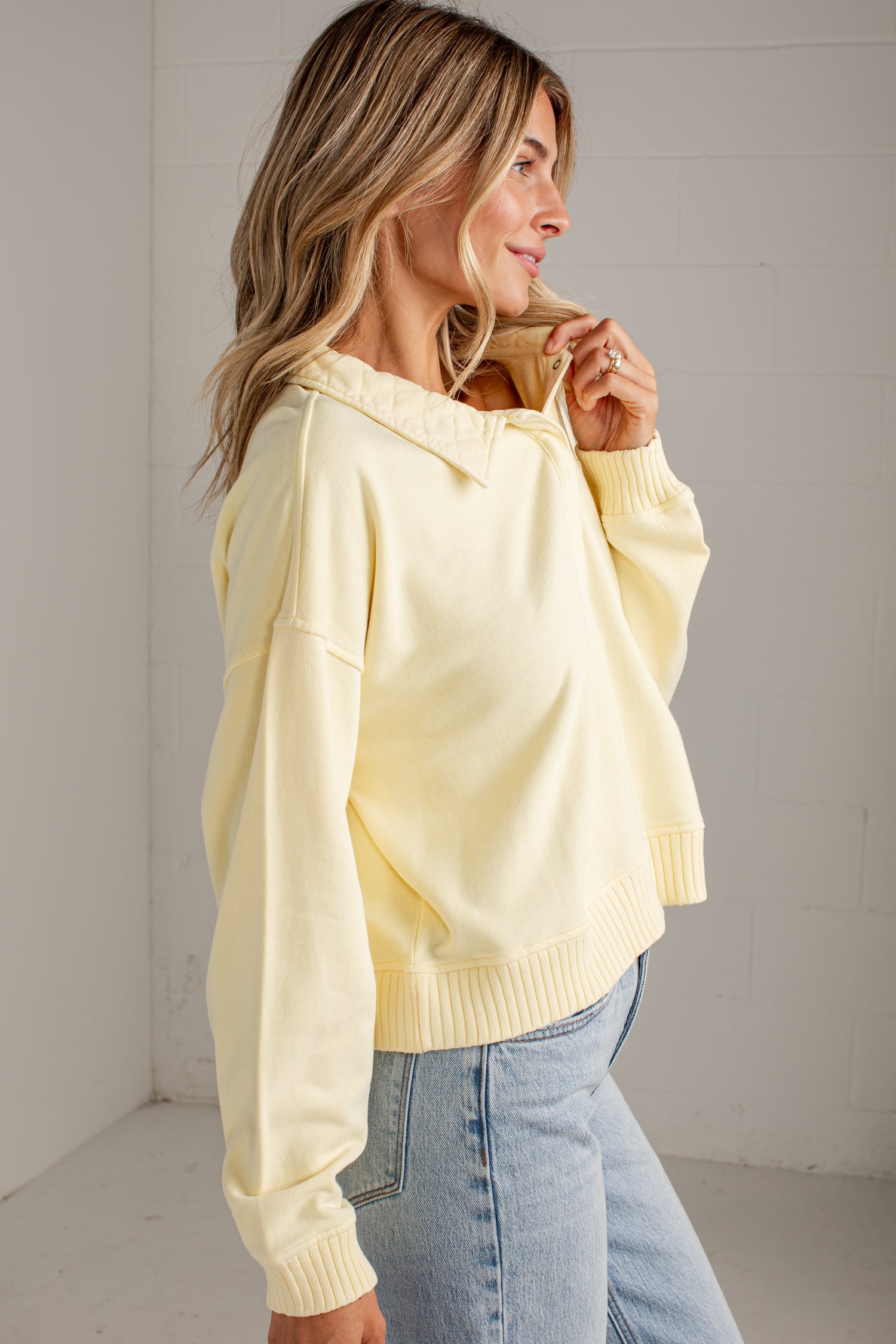 A woman wearing a pale yellow oversized knit sweater with a wide collar and ribbed cuffs, paired with light blue jeans, standing in a bright white room.