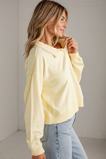 A woman wearing a pale yellow oversized knit sweater with a wide collar and ribbed cuffs, paired with light blue jeans, standing in a bright white room.