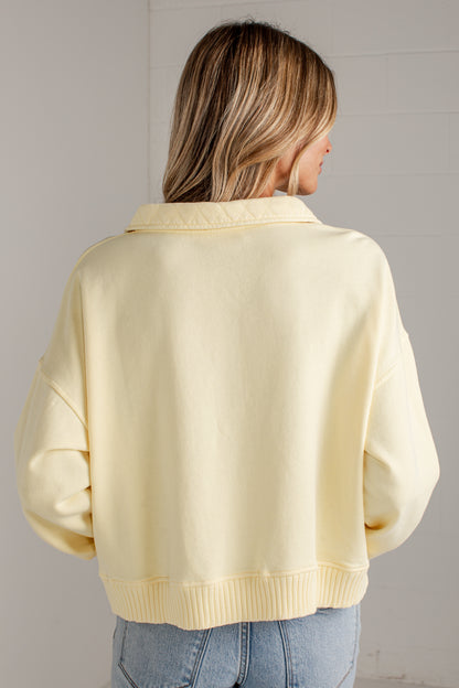 A woman viewed from behind wearing a pale pastel yellow cropped sweatshirt with a collar and ribbed hem, paired with light blue jeans against a light gray wall.