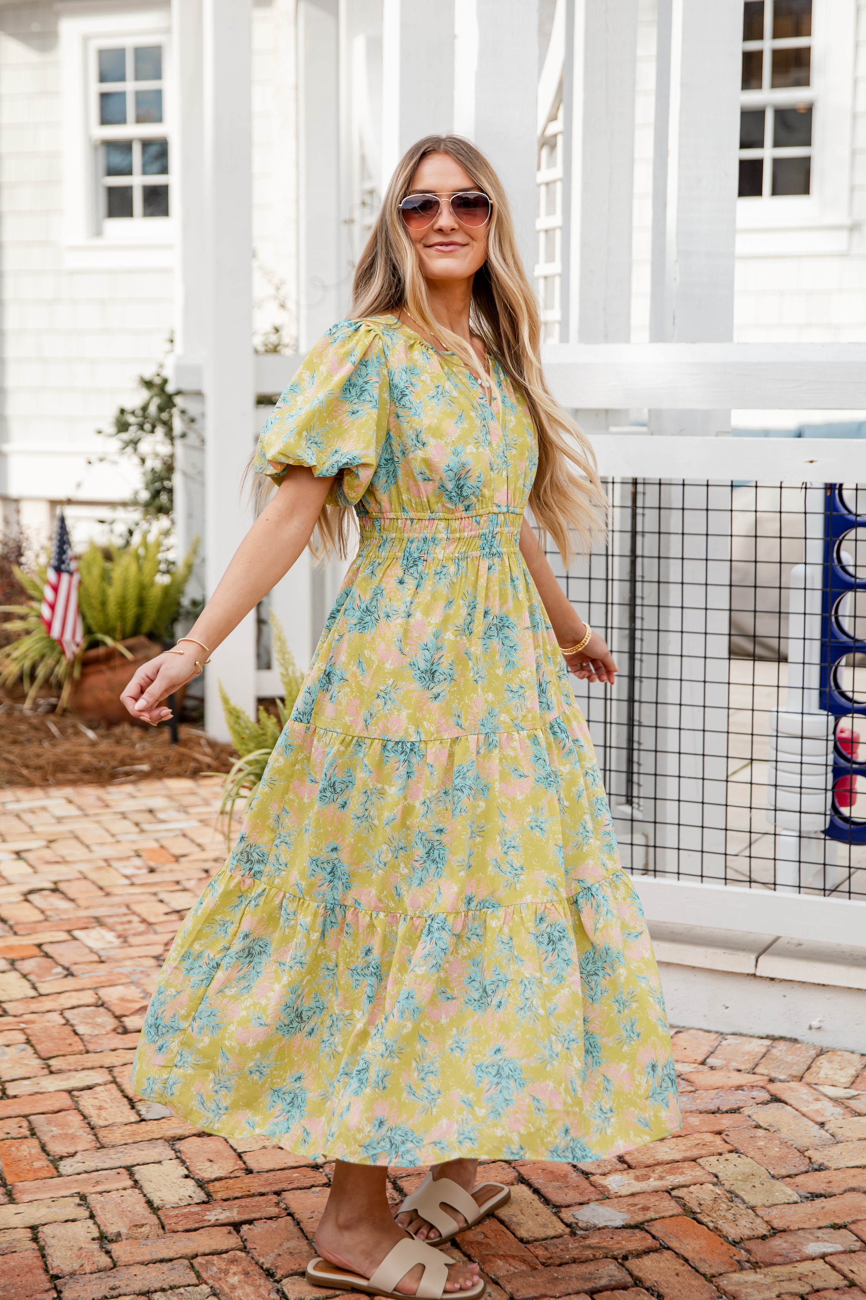 Woman wearing a yellow floral dress standing on a brick patio.