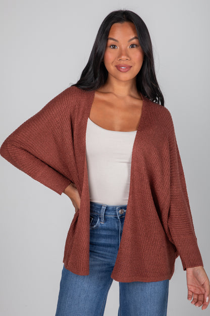 Woman wearing a brown cardigan over a white top and blue jeans on a gray background