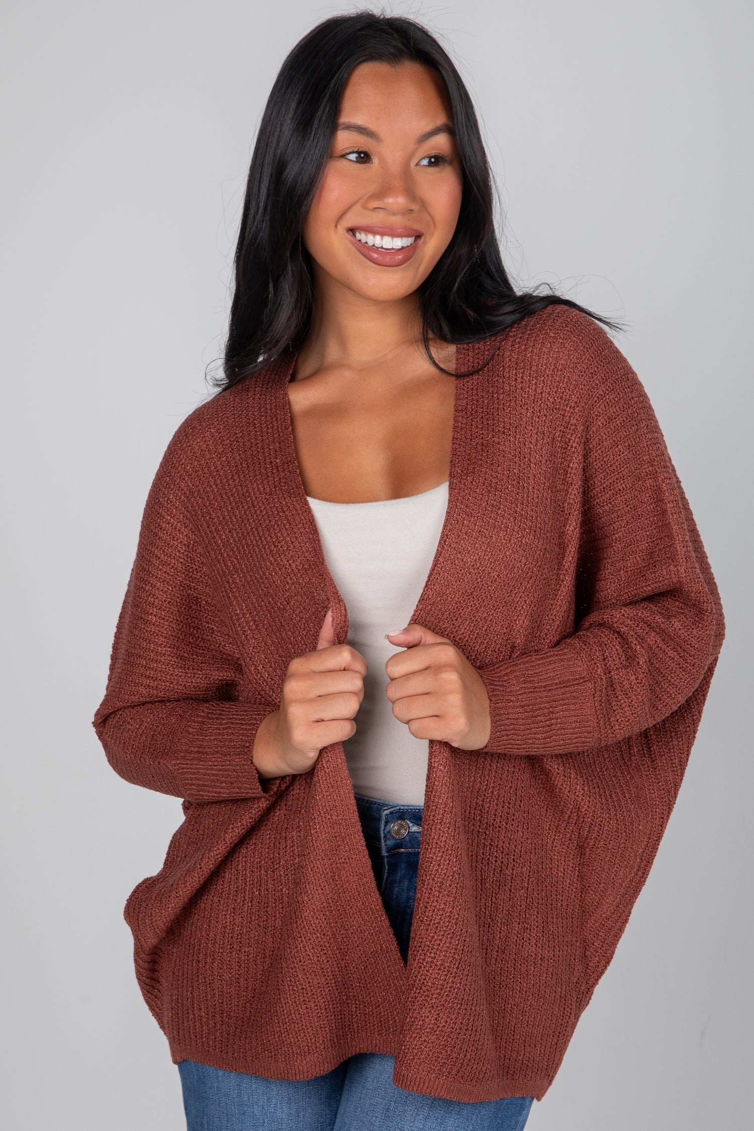 Woman wearing a brown cardigan over a white top and blue jeans on a gray background