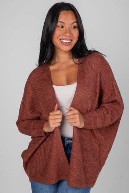 Woman wearing a brown cardigan over a white top and blue jeans on a gray background