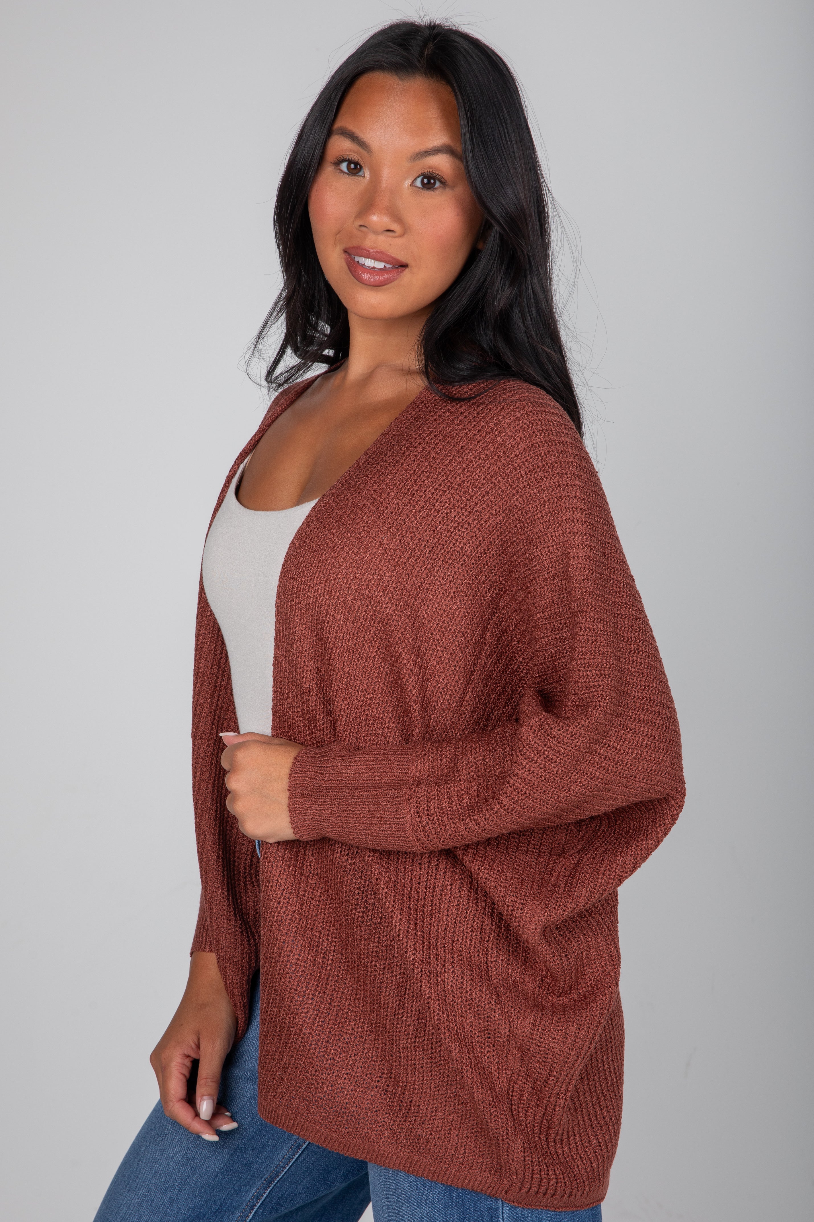 Woman wearing a brown cardigan over a white top and blue jeans on a gray background