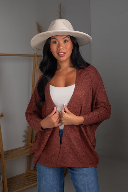Woman wearing a rust-colored cardigan, white top, blue jeans, and a beige hat indoors.