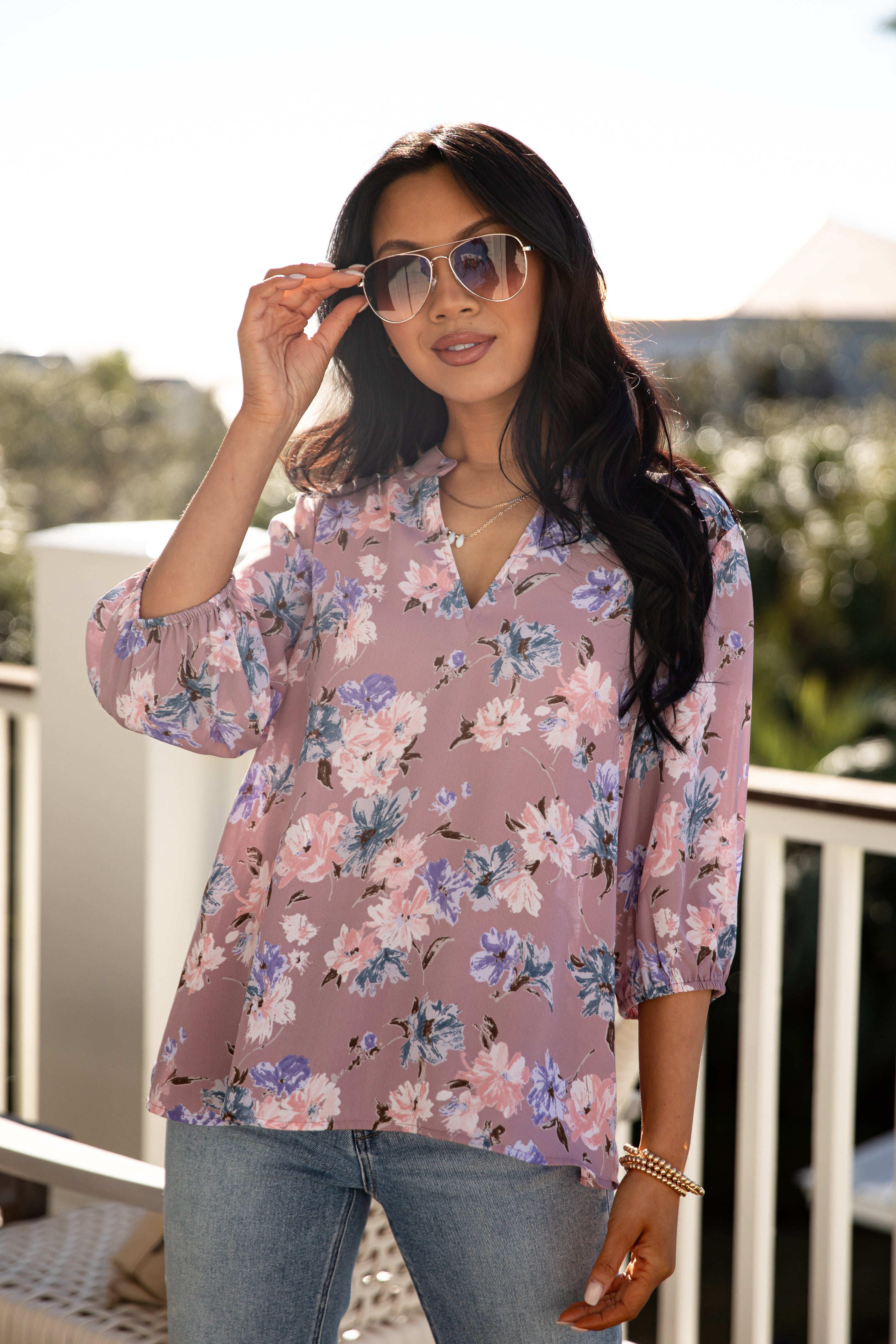 Woman wearing a floral blouse and sunglasses on a balcony with palm trees in the background