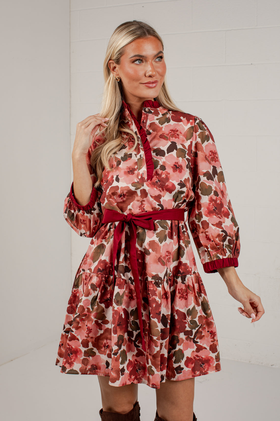 A woman modeling a long-sleeve floral dress with a red tie-neck collar, red waist tie belt, and red cuffed sleeves, set against a white brick wall background.