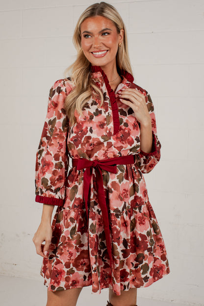 A smiling blonde woman models a short floral dress with a red collar, cuffs, and waist tie, set against a white brick wall.