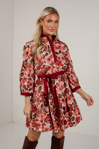 A woman modeling a floral long-sleeve mini dress with a matching red tie belt and red trim, paired with brown boots, standing against a white brick wall.
