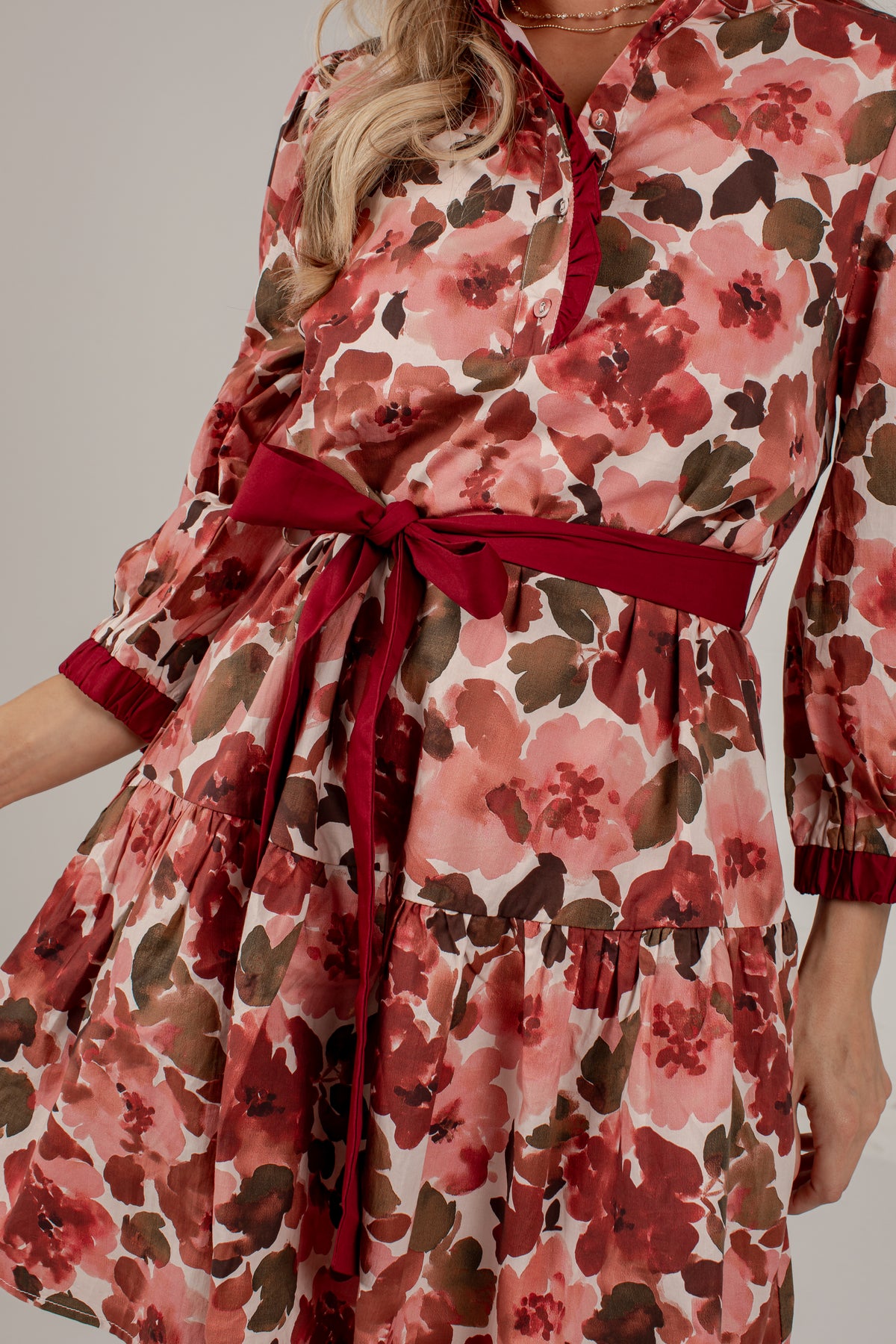 A close-up of a woman wearing a tiered floral dress with a red self-tie belt. The dress features a pink, red, and brown floral pattern on a cream background.