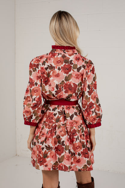 A woman viewed from behind wearing a floral long-sleeve mini dress with a red collar, red belt, and red cuffs, set against a white brick wall.