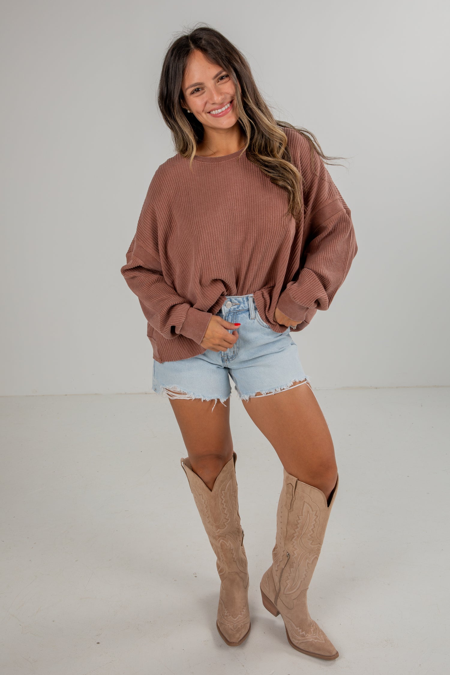 Woman wearing a brown sweater, light denim shorts, and tan knee-high boots on a white background