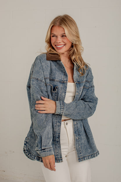 A smiling blonde woman wearing an oversized light-wash blue denim jacket with a brown collar, styled over a white tank top and white pants, standing against a white brick wall.