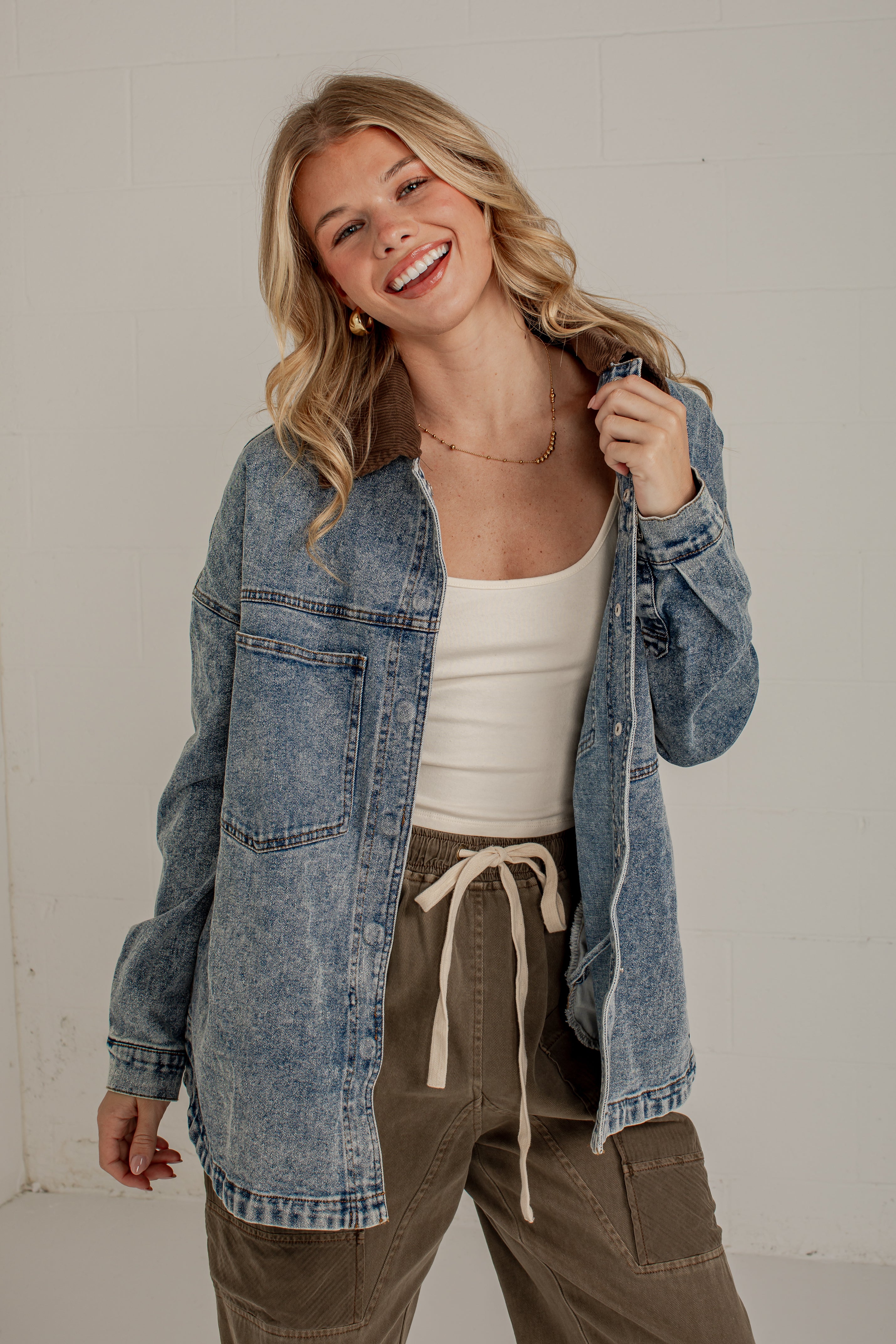 A woman smiling and wearing an oversized blue denim jacket over a white tank top and olive green drawstring pants, standing against a light gray wall.