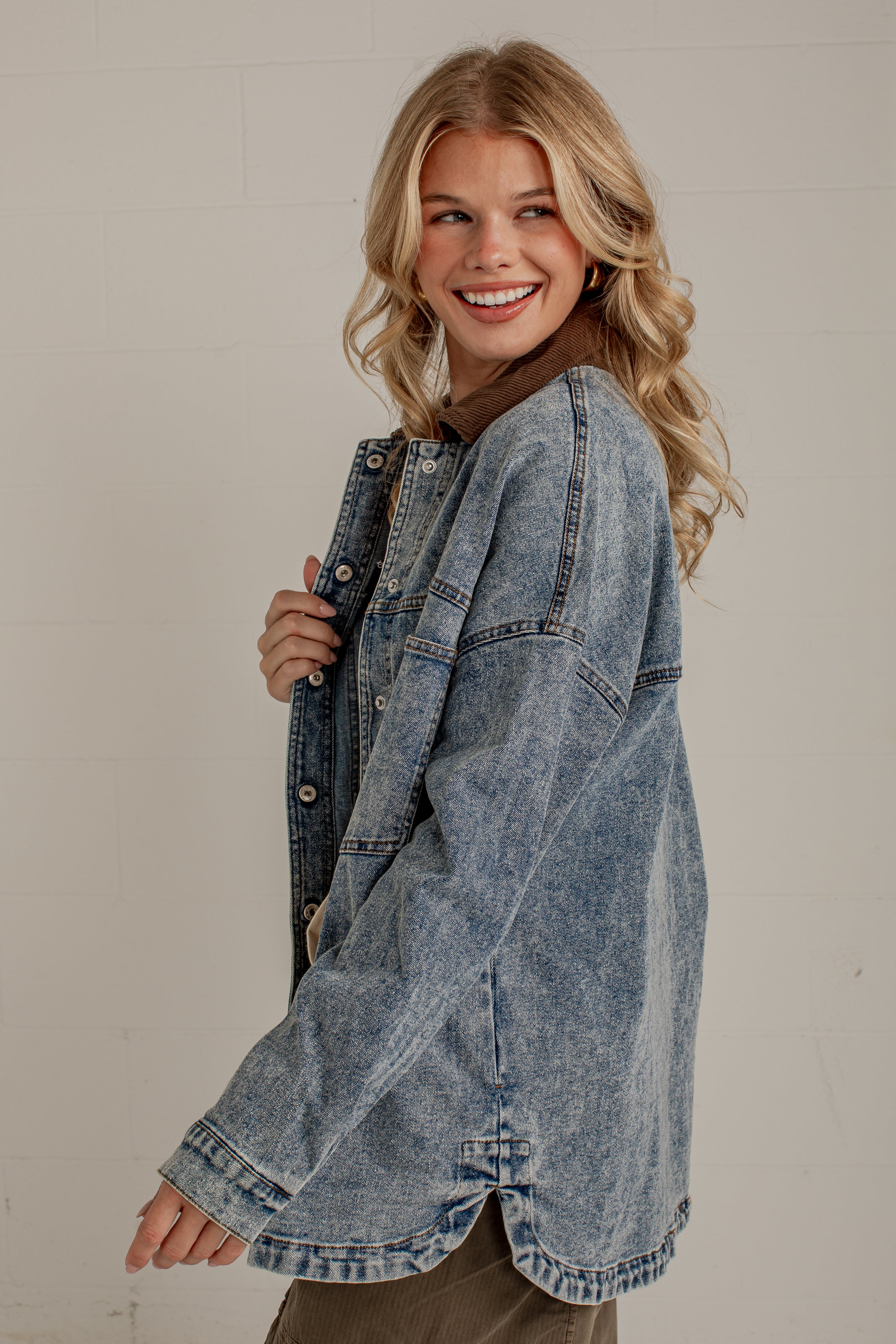A smiling blonde woman wearing an oversized light-wash blue denim jacket, posing against a light off-white brick wall.