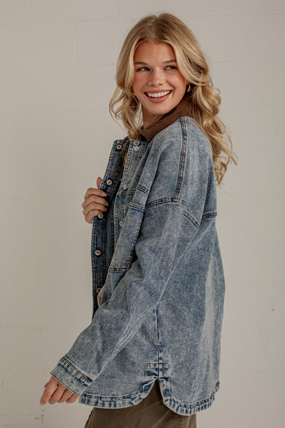 A smiling blonde woman wearing an oversized light-wash blue denim jacket, posing against a light off-white brick wall.